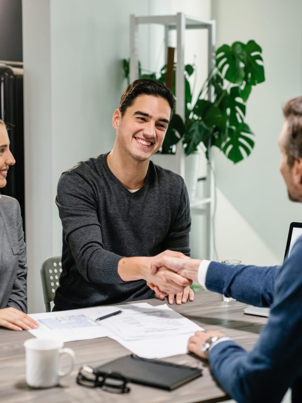 Happy couple shaking hands with their real estate agent at home. Young happy couple making an agreement with their financial advisor at home. Men are shaking hands.